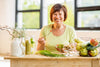 older lady smiling and sitting at a wooden table eating a healthy bowl of veggies