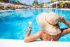 woman holding her sun hat and a drink in her hand while she stands in an outdoor pool.