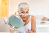 older woman looking in a small mirror as she applies under-eye masks.