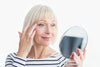 55-year-old woman applying skincare underneath her eyes to prevent dry skin, while looking in a small handheld mirror.