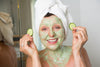 mature woman exfoliating with a green face scrub and cucumbers in both hands.