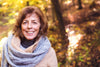 smiling senior woman, bundled up in a coat and scarf as she stands in the forest in the fall.