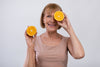 60-year-old woman holding up two halves of an orange - one is in front of her eye and the other is in her right hand.